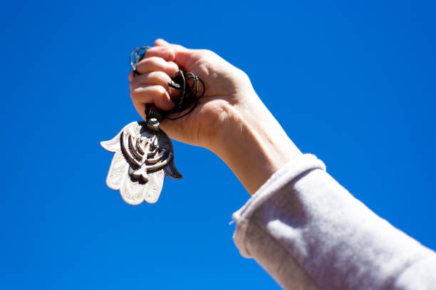 A hand holding up an antique (19th C) ornate silver Khamsa talisman pendant with Jewish menorah design against a vibrant blue sky. Originally from the Sephardic community in North Africa. Plenty of copy space on the blue background.
