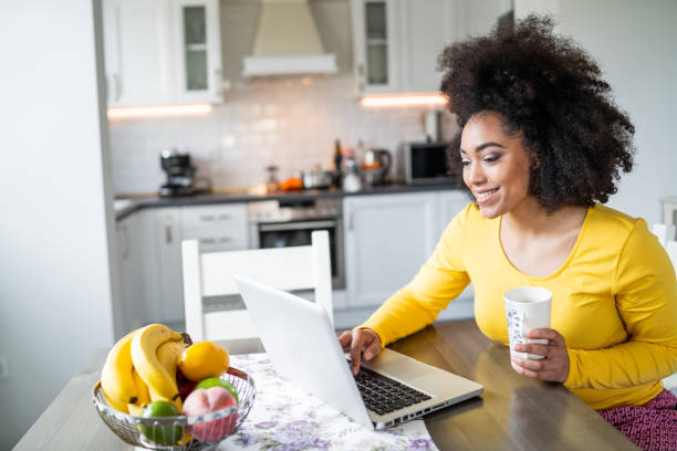 afro woman che usa il laptop a casa e beve caffè - working at home domestic kitchen laptop computer foto e immagini stock