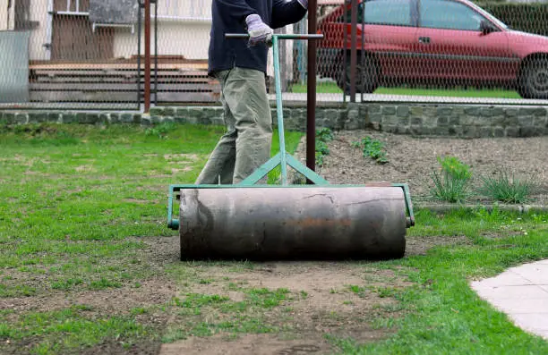 Photo of Summer worker pulls lawn roller for flatten garden. Heavy, iron cylinder tramples soil and then man can plant seeds. Important landscaping on garden in summer