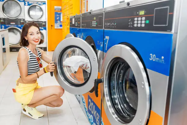 Young woman loading dirty clothes in washing machine in laundromat, laundry service concept