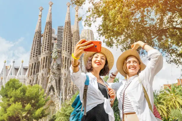 Photo of BARCELONA, SPAIN - 11 JULY 2018: Young girls friends making selfie photo on her smartphone in front of the famous Sagrada Familia catholic cathedral. Travel in Barcelona concept
