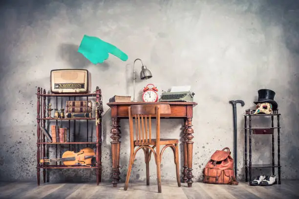 Photo of Vintage loft room with antique chair, old typewriter, lamp, clock on oak wooden desk, retro radio, books, fiddle, keys on shelf, mask, cylinder hat, shoes, cane, backpack, conceptual forefinger