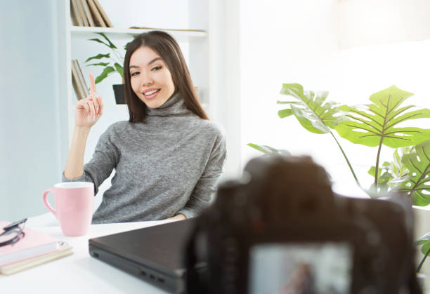 istock Beutiful blogger is sitting at the table and pointing up. She is recording on camera new video for her youtube account. 1144746632