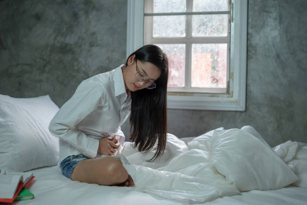 Young woman suffering from abdominal pain while sitting on bed at home, Hands of young woman on stomach from menstruation cramp, gastrointestinal, diarrheas or female diseases problem, healthcare concept. stock photo