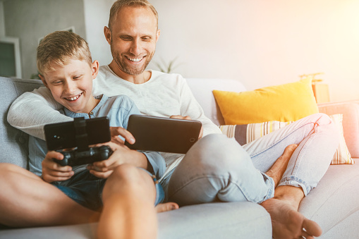 Father and son game players funs sit together at home on cozy sofa, using the tablet and gamepad