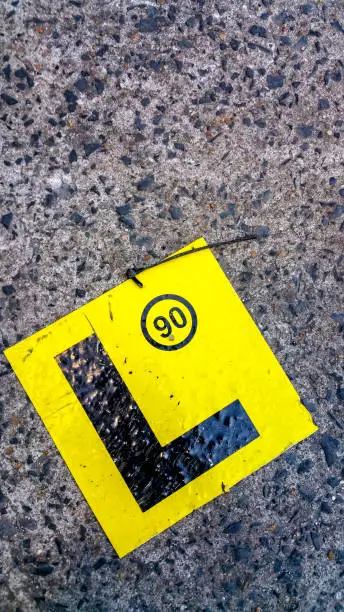A yellow motoring learner's L-Plate on a textured background. The "90" marking signifies the legally imposed speed limit in km/hr in which the driver can travel while in control of the vehicle.