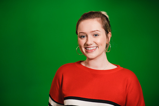 A front-view portrait shot of a young caucasian woman standing in front of a green screen, she is smiling and looking at the camera.