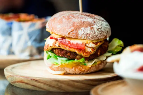 Close up image of a freshly flame grilled  vegetarian halloumi cheeseburger on a wooden counter at an outdoor food market. This burger is loaded with a vegetarian burger meat substitute, fresh salad, melted halloumi cheese and spanish onion and tomato. The burger is sandwiched between glazed buns. Horizontal colour image with copy space.