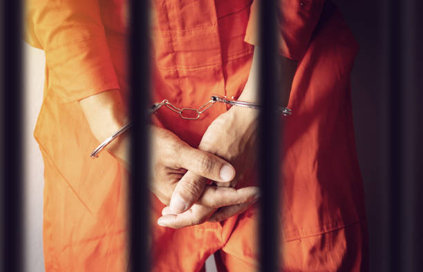 un prisionero con las esposas detrás de los barrotes de una prisión de ropa de mono naranja - orange uniform fotografías e imágenes de stock