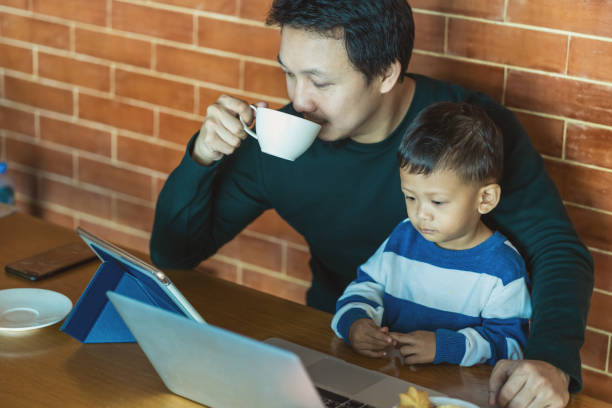 papa célibataire asiatique avec le fils regardent le dessin animé par l’ordinateur portatif de technologie et le café potable avec le travail quand vivant dans la maison de loft pour l’auto-apprentissage ou l’école à la maison, la famille et le  - brown boy photos et images de collection