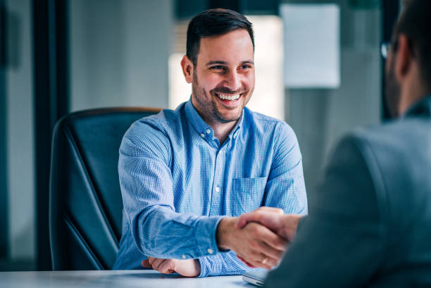 two smiling businessmen shaking hands while sitting at the office desk. - sold imagens e fotografias de stock