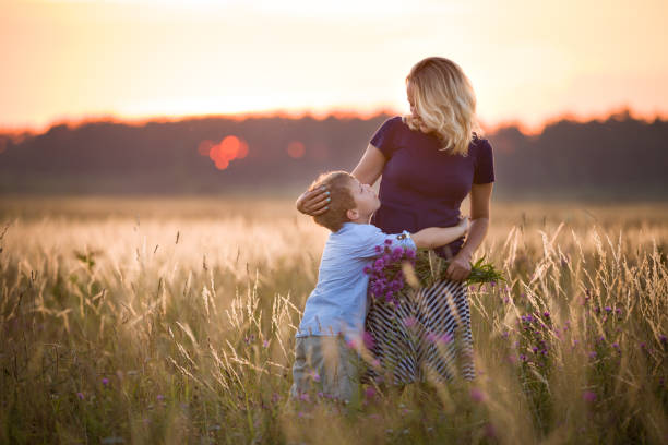 carino ragazzo ragazzo che abbraccia sua madre su un prato estivo sul bellissimo tramonto estivo. famiglia felice insieme. mamma e figlio. maternità e infanzia. famiglia che cammina sul campo. fuori. - natura di madre foto e immagini stock