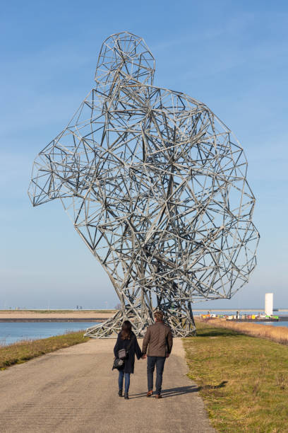 Lelystad, The Netherlands - February 23, 2019: Walking couple near iron statue of a man sitting on his heels on an enclosure dam in Lelystad, The Netherlands