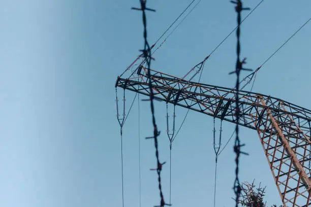 High voltage electricity pole from low angle against clear blue sky with barbed wire in foreground, protecting power line
