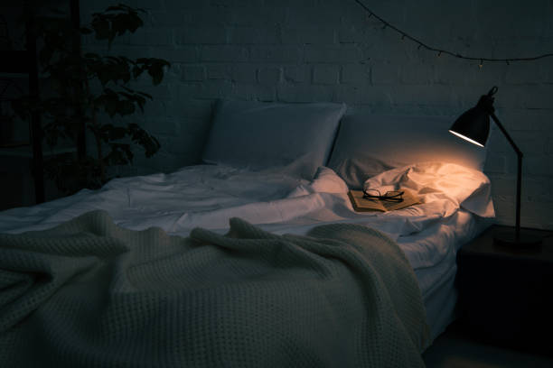 Interior of bedroom with book and glasses on empty bed, plant and lamp on black nightstand at night Interior of bedroom with book and glasses on empty bed, plant and lamp on black nightstand at night night table stock pictures, royalty-free photos & images