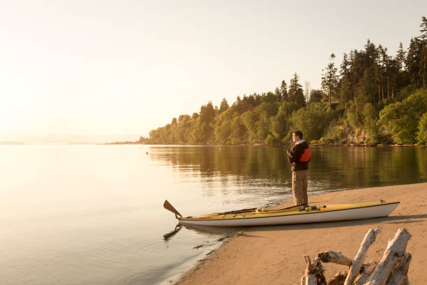 man with kayak on beach looking out at water. solo outdoor adventure sports sea kayaking in beautiful, remote nature wilderness. - langley imagens e fotografias de stock