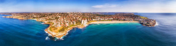 D Sy Bondi Sculpture High Pan Famous Bondi beach on Sydney Pacific coast in elevated aerial wide panorama between sandstone headlands with distant city CBD on the horizon. bondi beach photos stock pictures, royalty-free photos & images