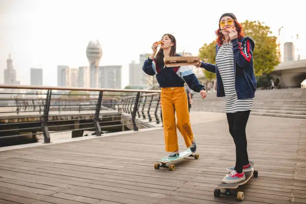 Red hair woman and her Asian female  friend having fun  while riding long boards and eating pizza