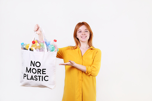 Emotional redhead woman holding eco friendly shopping bag with no more plastic text, full of harmful products, bottles and bags. Zero waste concept. Happy female smiling, separating garabage.
