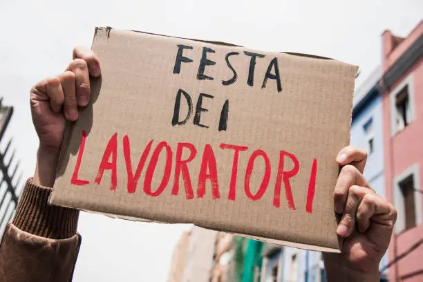 closeup of a young caucasian man outdoors showing a brown cardboard signboard with the text festa dei lavoratori, may day in italian, handwritten in it