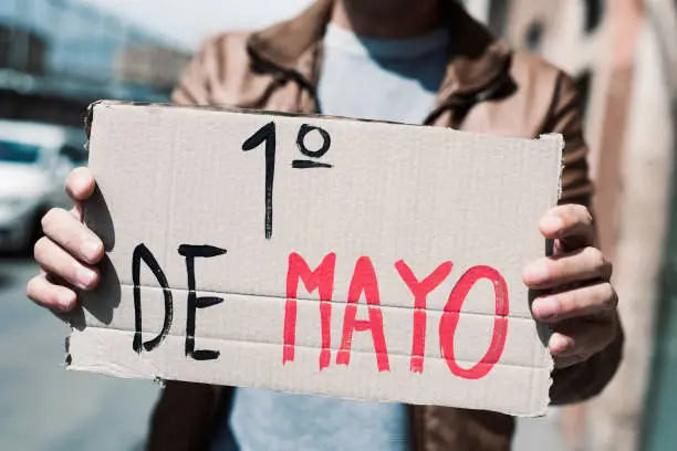 closeup of a young caucasian man outdoors showing a brown cardboard signboard with the text 1 de mayo, may day in spanish, handwritten in it