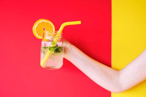 Woman hand holding Summer cocktail on multi colored background, cocktail with mint and orange fruit with ice.