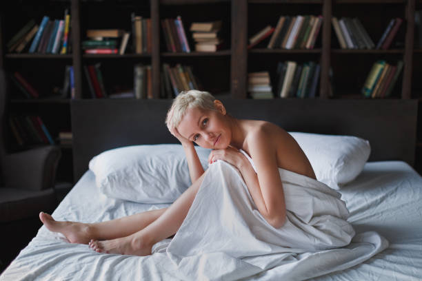 Pensive beautiful young woman with blond short hair sitting on bed on white linen stock photo