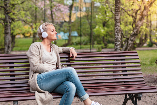 A middle-aged woman is listening to music through a pair of white headphones. She is sitting on a bench in a park with trees around her.