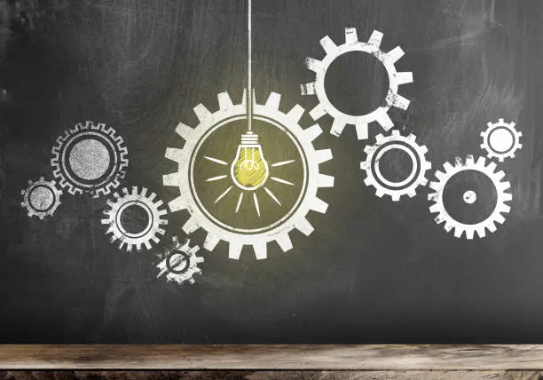 Photo of gear with interlocking cogwheels with glowing light bulb on blackboard