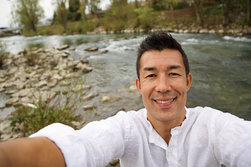 Mid-adult man making selfie with river as the background. Casual clothes, happines on his face. Enjoying the day.