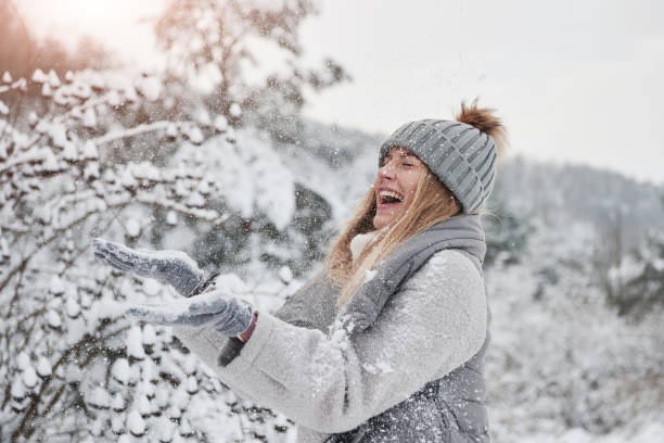sorridere e divertirsi. ragazza allegra in abiti caldi che gettano neve all'aperto vicino alla bellissima foresta - snow heat adult blond hair foto e immagini stock