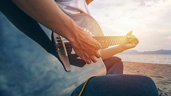 man's hands playing acoustic guitar, capture chords by finger on sandy beach at sunset time. playing music concept