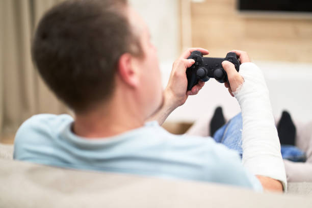 istock Man with broken arm in plaster cast holding controller and playing in videogame in front of TV. Game addiction concept. 1145025995