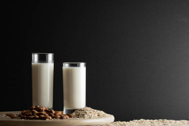 Two glasses of oat milk and almond milk with oat flakes and almond nuts on wooden disk. Copy space, black background. stock photo