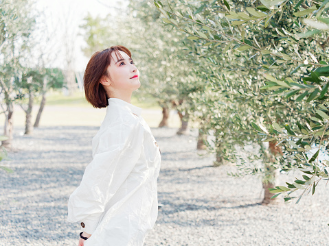 Portrait of a beautiful Chinese woman in white shirt stand in orchard in sunny day, looking upwards with hand behind herself, elegant girl.