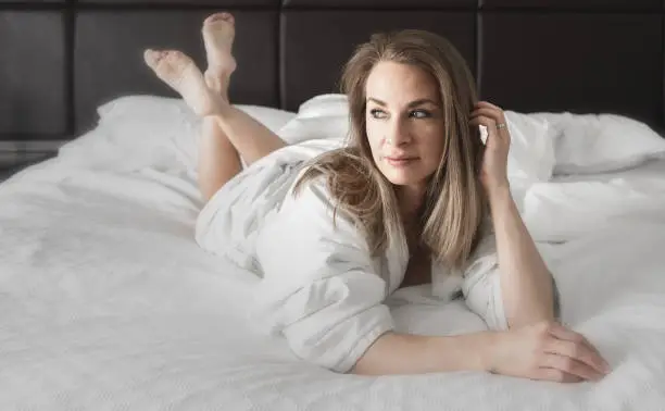 Nice looking brunette woman in white bathrobe relaxing in her white linen bed in the morning. Soft focus brunette woman.
