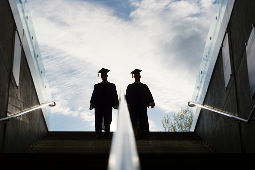 This is the silhouette of two college graduates in cap and gown climbing up a set of steps.  This shot is backlit with bright morning sunshine in the background.