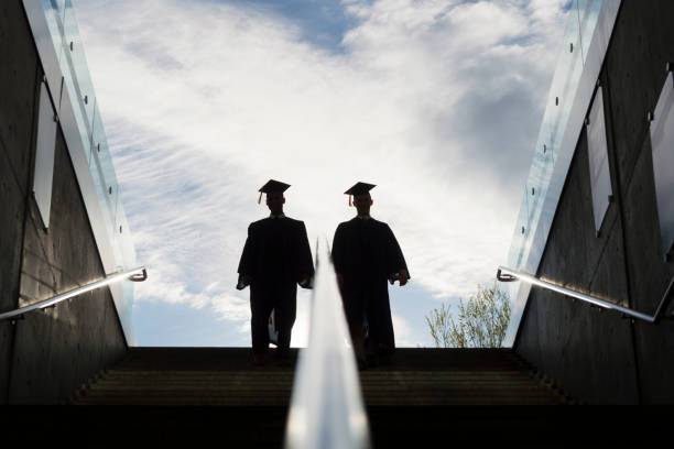 Silhouette of Two College Graduates Climbing Steps This is the silhouette of two college graduates in cap and gown climbing up a set of steps.  This shot is backlit with bright morning sunshine in the background. continuing education stock pictures, royalty-free photos & images