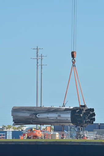 A rocket booster from a SpaceX falcon 9 heavy launch is hoisted by crane at the dock adjacent to a barge. It is being prepared for land transport. The center booster rocket was damaged after landing on the barge. SpaceX hopes to reuse the rocket engines.