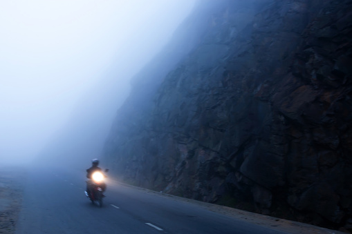 An unidentified male riding motorcycle on the dark misty mountain road at dusk, mystic asphalt road in mountain gorge with light trail from headlight leading through winter foggy. Motion blur.