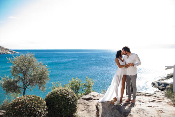 Man holding woman, his arms wrapped around her waist and she is holding on to him, standing in a seascape near the mediterranean sea. stock photo