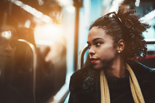The portrait of a young charming African-American female pensively looking outside the carriage window while sitting indoors of a metro train; Brazilian girl in a subway train, shallow depth of field