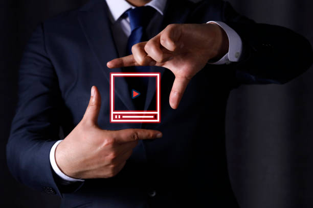 A businessman in a navy suit and blue tie gestures with his hands to frame a virtual video player icon with a play button, set against a dark background.