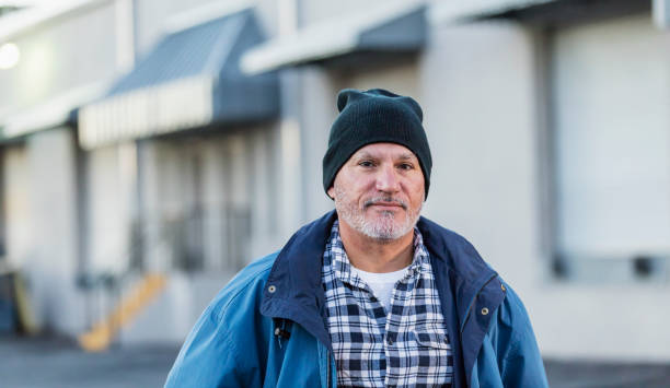 Mature Hispanic man standing outside warehouse A mature Hispanic man with a beard, in his 50s, standing outside a warehouse wearing a coat, plaid shirt and knit hat, looking at the camera. man beard plaid shirt stock pictures, royalty-free photos & images