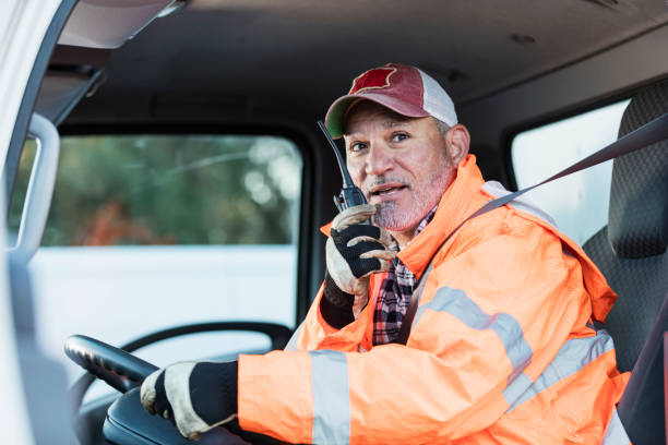 Hispanic man driving a truck A mature Hispanic man in his 50s, a truck driver, is on his walkie-talkie, sitting in the driver's seat. He is wearing a trucker's hat, reflective jacket and protective gloves. one mature man only audio stock pictures, royalty-free photos & images