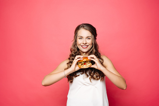 Close up of hungry girl with opened mouth, holding and eating big hamburger. Pretty woman with curly hairstyle and red lips eating tasty cheeseburger with tomato and meal. Concept of fast food.