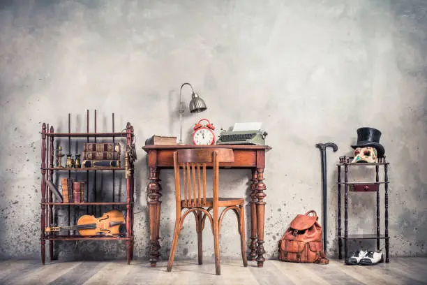 Photo of Room interior with antique chair, old typewriter, lamp, retro clock on wooden desk, books, binoculars, fiddle, keys on shelf, mask, cylinder hat, shoes, cane, backpack. Vintage style filtered photo