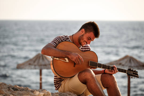Happy young man sitting on sand and playing an acoustic guitar at the beach during sunny summer day.