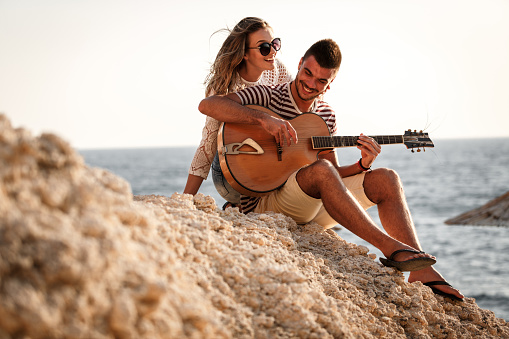 Young man playing an acoustic guitar while relaxing with his girlfriend by the sea.
