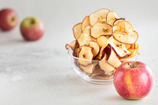 istock Hipped Dried apple chips in glass bowl with fresh apple in foreground on light background - Image 1145125003